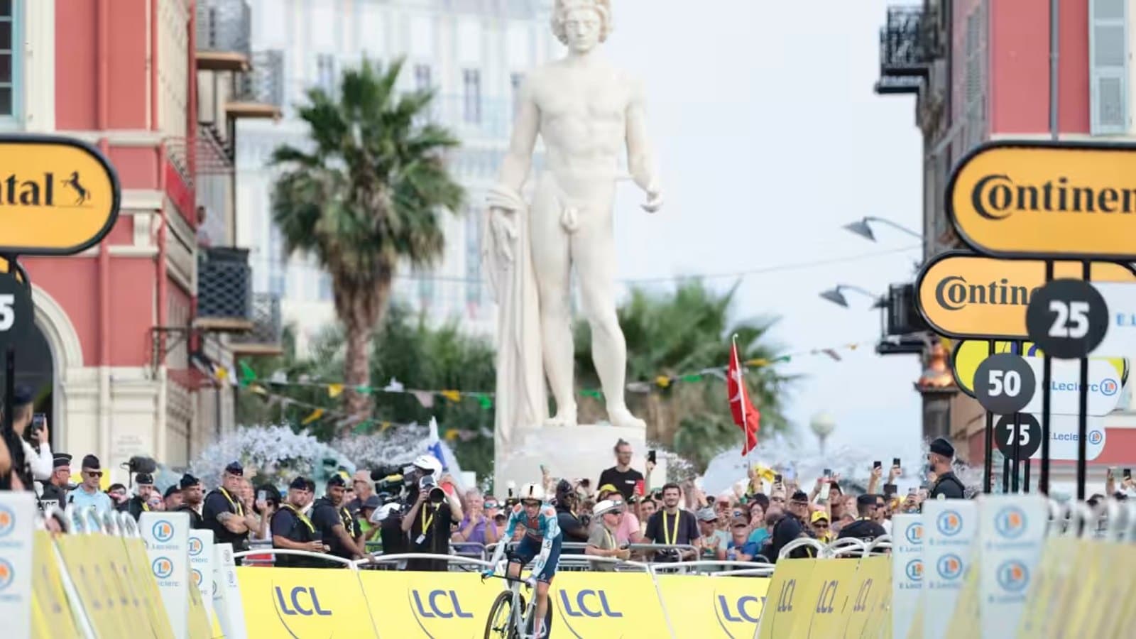 A cyclist racing past a large statue and crowd barriers marked with kilometre countdowns and sponsor branding near a Grand Tour finish line.