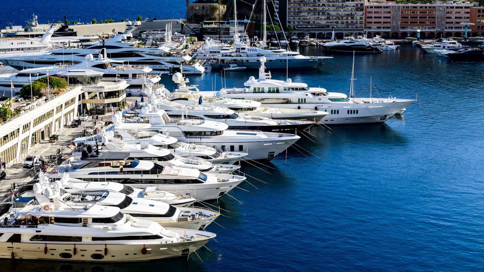 Rows of superyachts moored at Port Hercule in Monaco, with the harbourfront buildings behind them.