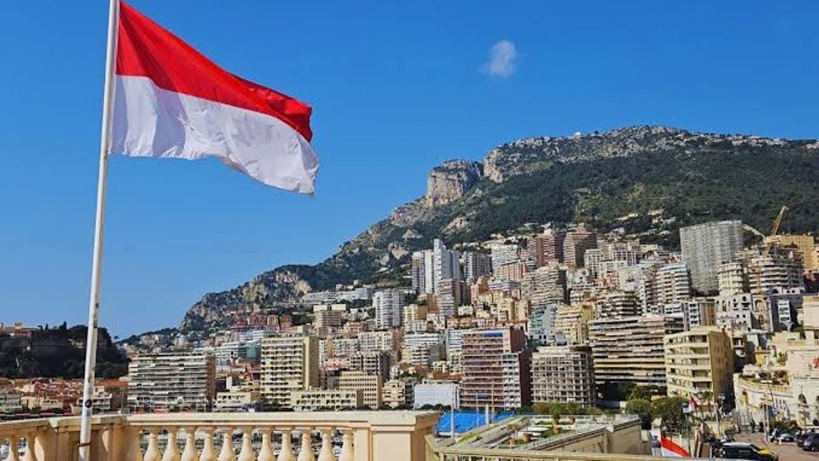 The Monegasque flag flying above the rooftops of Monaco, with Monte Carlo's hillside towers and the Tete de Chien cliff in the background.