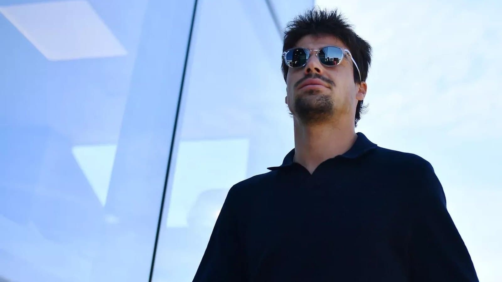 A portrait of Formula 1 driver Lance Stroll wearing sunglasses and a dark polo shirt, photographed from below against a glass building and blue sky.