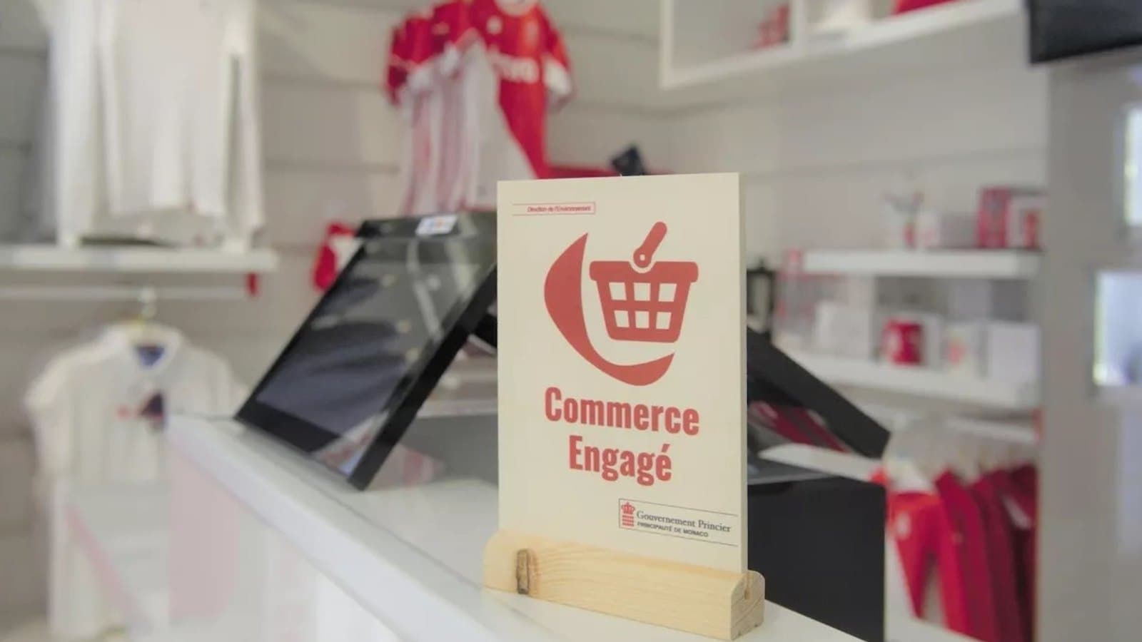 A Commerce Engagé placard on a shop counter, reading Commerce Engage above the crest of the Gouvernement Princier, with red and white merchandise on the shelves behind it.