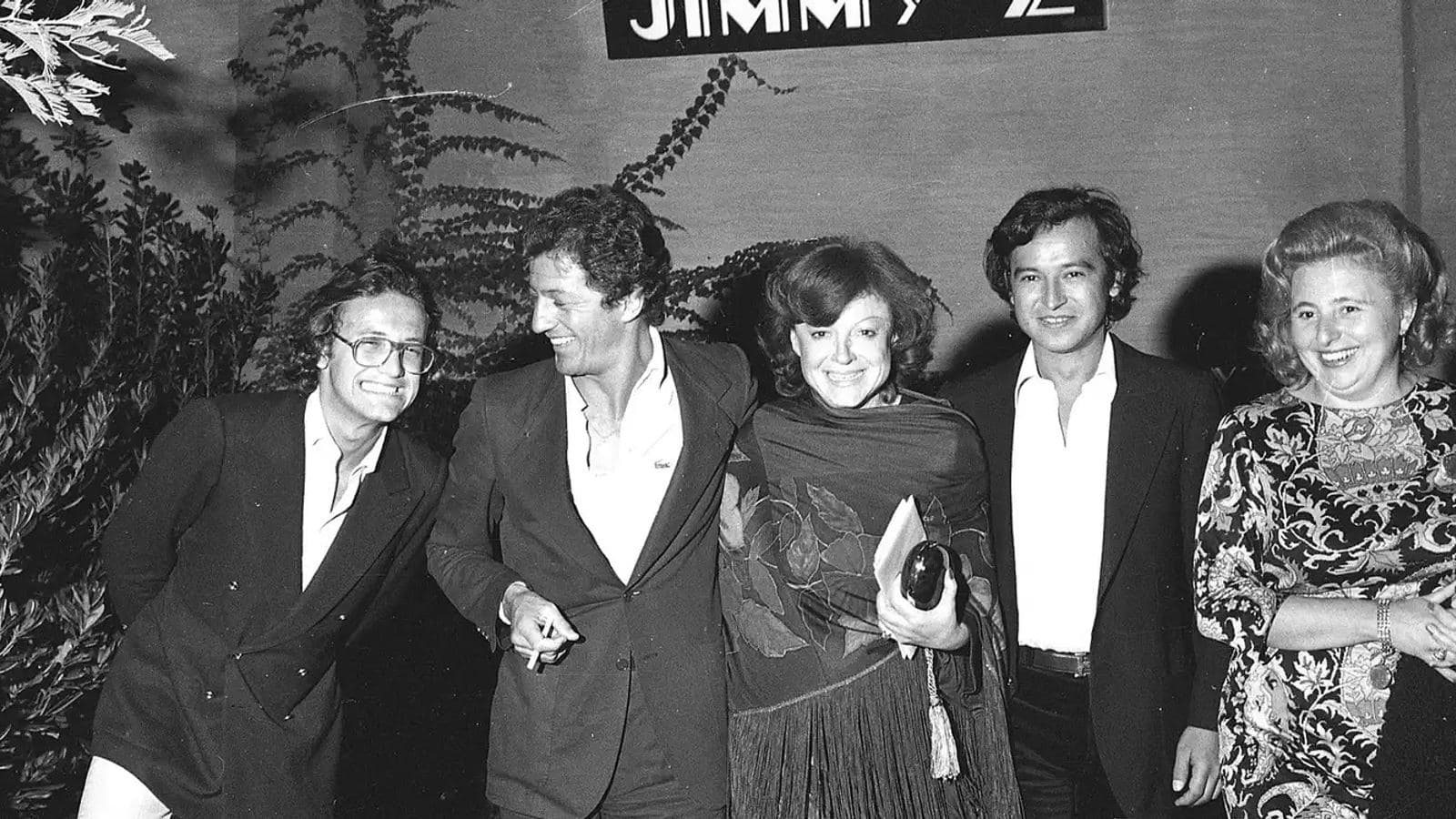 A black and white archive photograph of five smiling guests posing together beneath the Jimmy'z sign at the club in Monte Carlo.
