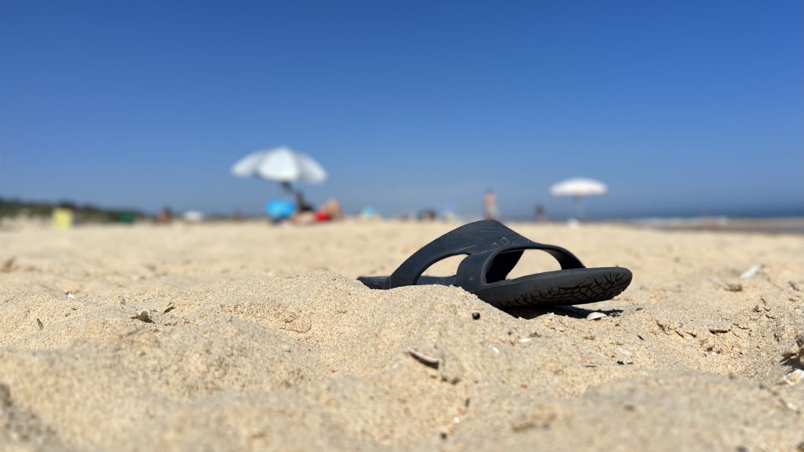 Beach. Close up view of the ground. Just over the top half of the photo shows the clear blue sky in the background, and below, blurred, some parasols and a few people. In the lower part, a dark blue flip flop stands out on the light sand.