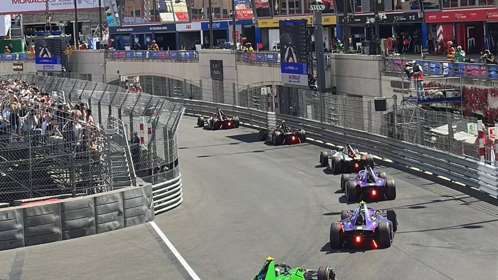Formula E cars racing through Monaco's Circuit de Monaco during the Monaco E-Prix, with the crowd watching from trackside stands.