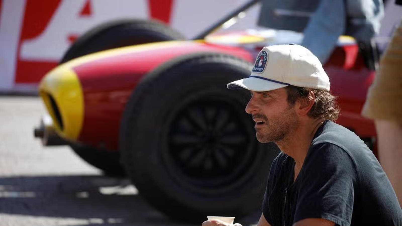 Bradley Cooper sitting beside a vintage Formula 1 car on set in Monaco, wearing a cap and holding a coffee cup.