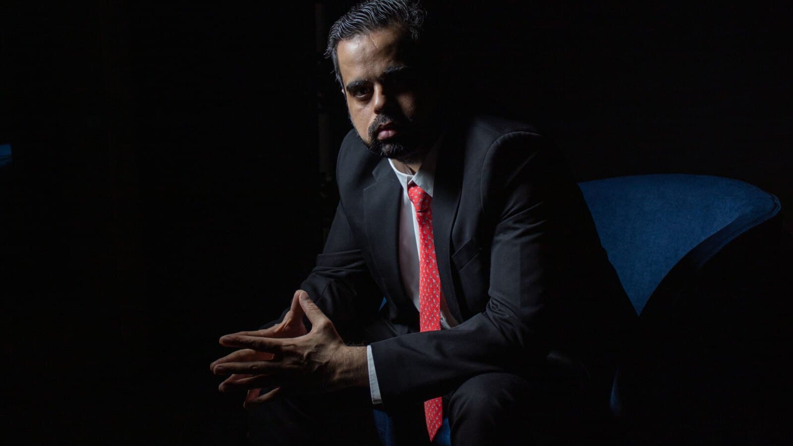 A portrait of businessman Prateek Suri seated in a dark room, wearing a black suit and red tie.