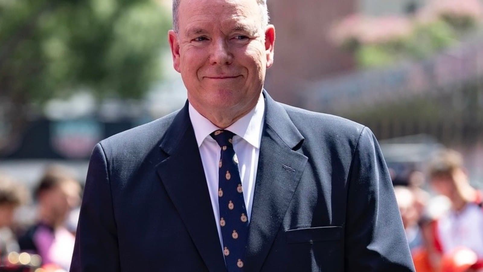 A portrait of Prince Albert II of Monaco, smiling, wearing a dark suit and a tie with the Monaco palace crest.