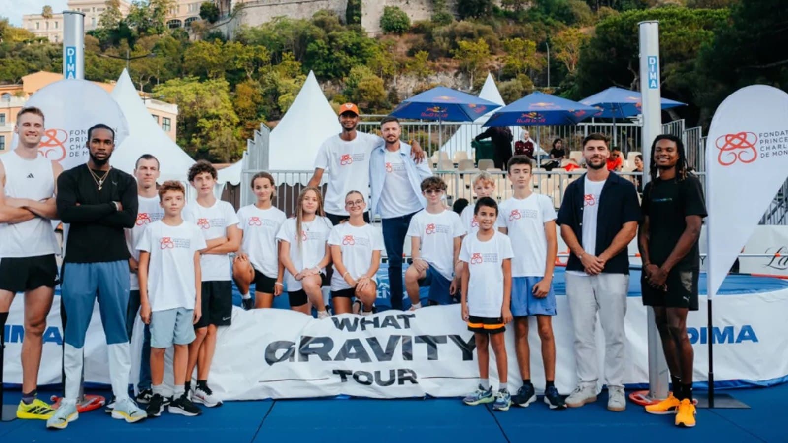A group of professional athletes and young club members posing behind a What Gravity Tour banner, with the Fondation Princesse Charlene de Monaco logo on their t-shirts and the Rock of Monaco in the background.