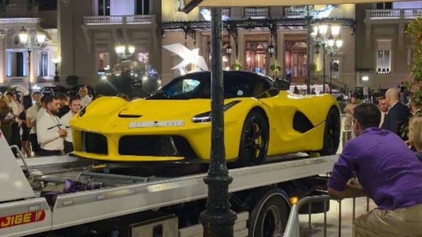 A yellow Ferrari LaFerrari loaded onto a flatbed recovery truck in Casino Square, Monaco, with a crowd gathered around the Casino de Monte-Carlo at night.