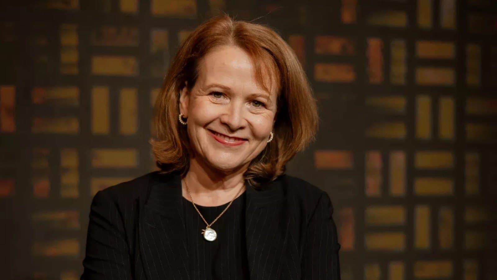 A portrait of a smiling woman with shoulder length auburn hair, wearing a black pinstripe blazer and a gold pendant necklace.