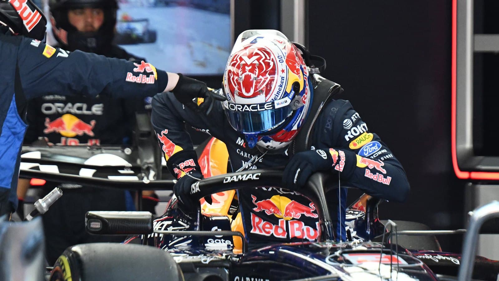 Max Verstappen climbing into his Red Bull Formula 1 car in the garage, wearing his helmet, with team mechanics beside him.