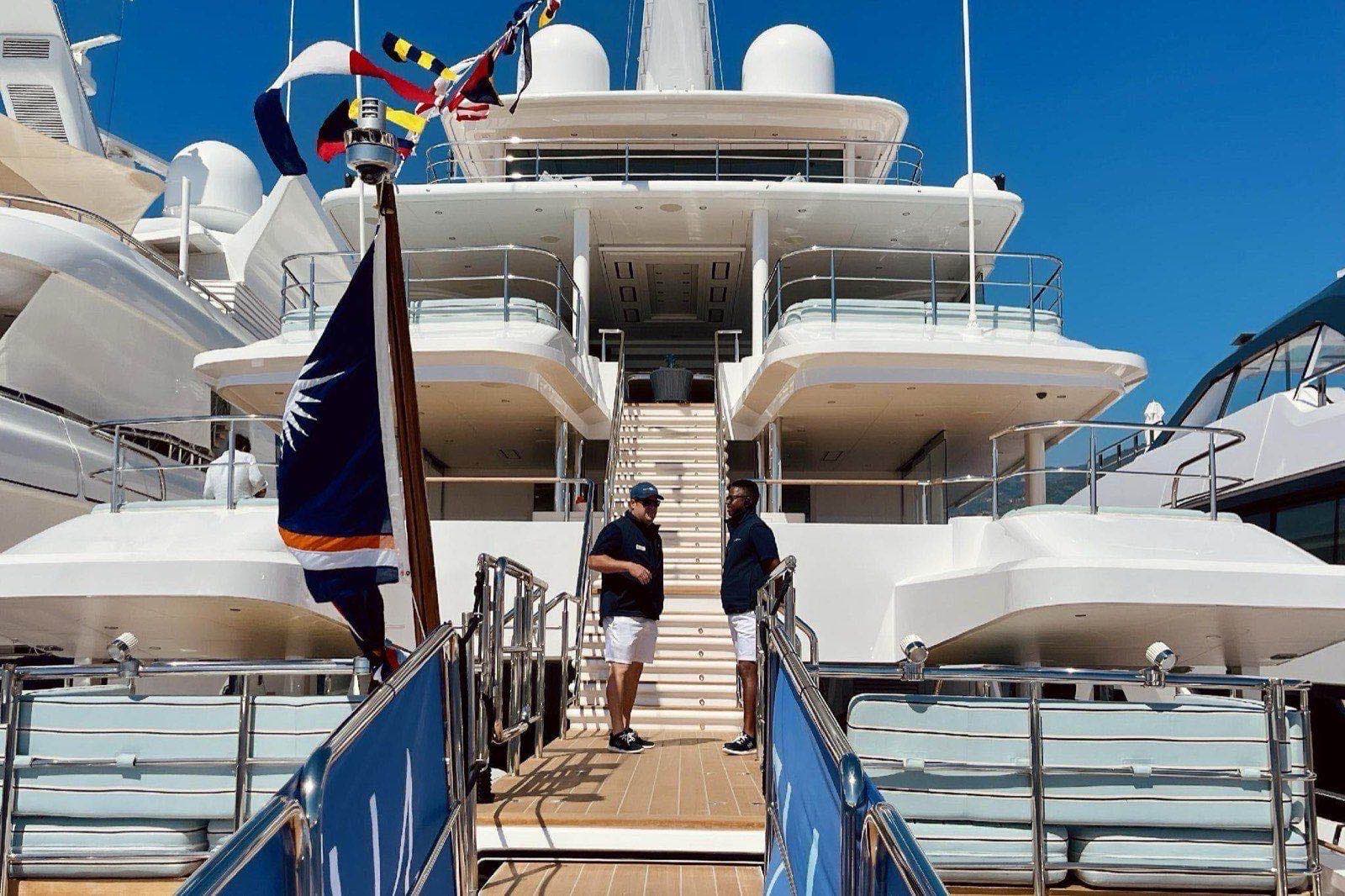 Two crew members in navy polos stand on the stern platform of a superyacht flying signal flags, with other yachts moored alongside in the marina.
