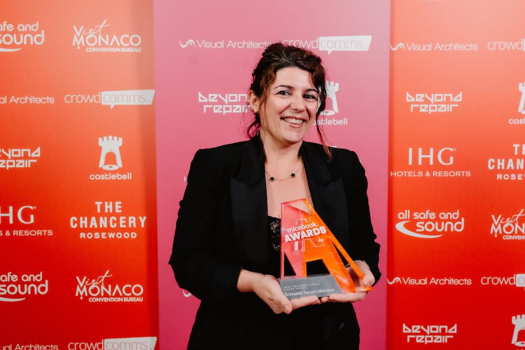 Une lauréate sourit en tenant son trophée orange aux micebook Awards, devant un mur de logos de sponsors